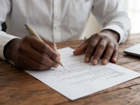 Une main signe un document juridique sur un bureau en bois avec lumière naturelle.
