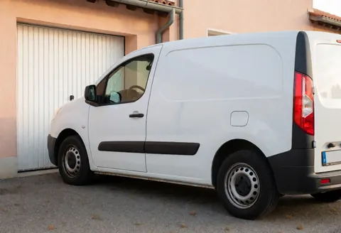 Une camionnette blanche est stationnée devant un garage avec des murs en terracota pâle, sous une lumière douce de fin de journée, évoquant les services de terrain.