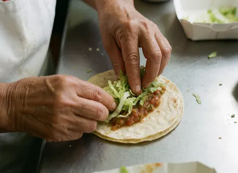 Des mains préparent un taco sur un plan de travail métallique brillant, avec des ingrédients frais et une texture croustillante visible.