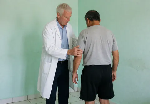 Un professionnel en blouse blanche examine le bras d'un patient dos à la caméra dans un cabinet aux murs verts et au sol blanc.