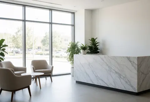 Réception de bureau moderne avec un comptoir en marbre blanc, des chaises confortables et des plantes vertes. Grandes fenêtres laissant entrer la lumière naturelle. Ambiance propre, minimaliste et accueillante.