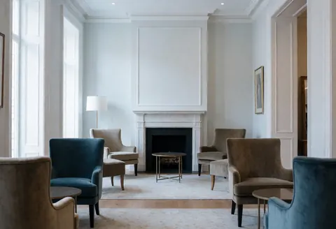 Vue d'un hall d'hôtel luxueux et silencieux baigné de lumière matinale, avec des fauteuils en velours, une cheminée et de grandes fenêtres.