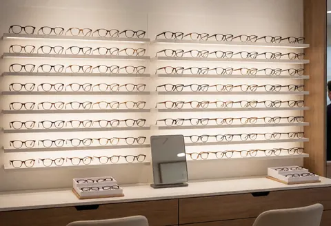Mur présentant des lunettes dans une boutique d'optique, éclairage doux, comptoir propre, miroir et présentoirs contenant des lunettes.