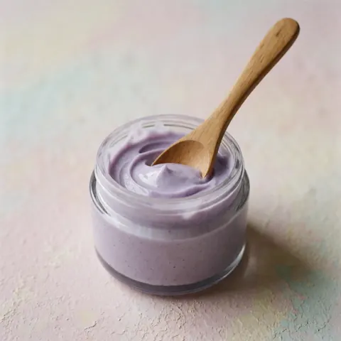 Pot en verre contenant une crème de lavande avec une cuillère en bois sur un fond pastel doux et éclairage naturel.