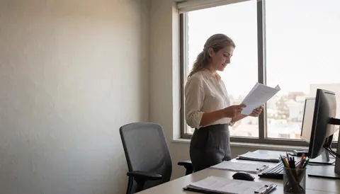 Une professionnelle vérifie des papiers sur un bureau équipé d'un ordinateur, près d'une fenêtre laissant entrer la lumière naturelle.