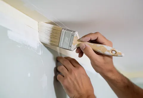 Main tenant un pinceur près d'un mur avec du ruban de masquage, peinture éclatante et propre, plafond et mur visibles, éclairage doux et professionnel.
