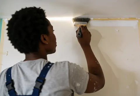 Une peintre professionnelle applique de la peinture blanche sur un mur intérieur avec un pinceau, dans un environnement de travail réel.