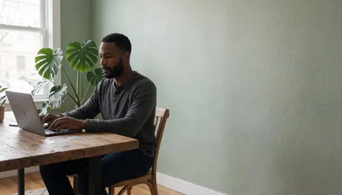 Un agent professionnel travaille sur un ordinateur portable posé sur un bureau en bois récupéré, dans un espace de travail lumineux avec une plante verte et un mur vert sage.