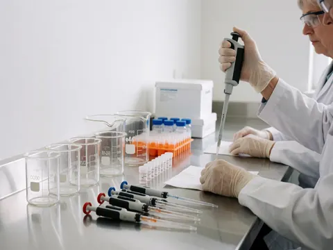 Un professionnel du laboratoire clinique manipule des pipettes et un flacon d'échantillon, portant des gants et des lunettes de protection sur une table en métal brillant.