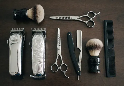 Vue de dessus d'outils de barbier incluant ciseaux, tondeuse, peigne et brosse posés sur une surface en bois foncé avec un éclairage doux.