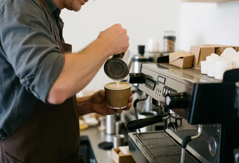 Un barista prépare un café latte en versant du lait dans une tasse, mettant en valeur l'art du latte et la qualité du service en cafétéria.