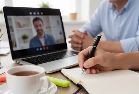 Une personne écrit dans un carnet pendant une visioconférence, avec un ordinateur portable, une tasse de thé et un marqueur sur le bureau.