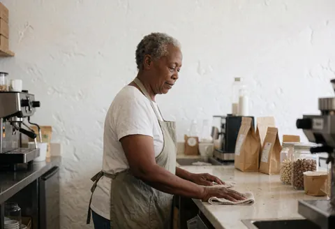 Une femme âgée portant un tablier essuie le comptoir avec une serviette dans un café aux murs blancs, machines et sacs de café visibles.