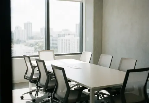 Salle de réunion moderne avec table blanche, chaises ergonomiques, grande fenêtre vue ville, murs béton, lumière naturelle, ambiance professionnelle.