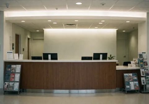 Intérieur d'une clinique médicale moderne montrant un bureau d'accueil en bois, des écrans informatiques, des brochures et une lumière douce. Ambiance professionnelle et propre.