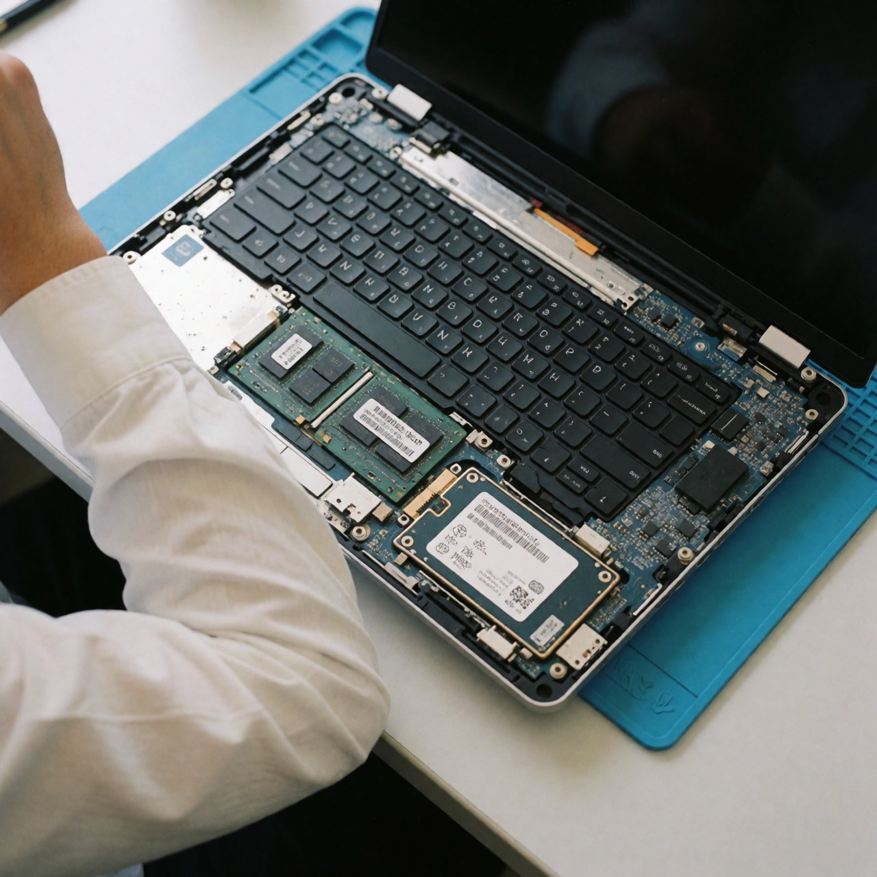 Laptop ouvert montrant la RAM et le SSD installés, main d'un technicien travaillant sur un tapis antistatique bleu dans une scène professionnelle de réparation informatique.