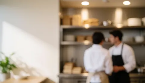 Cuisine de restaurant avec personnel en arrière-plan, étagères en acier inoxydable et éclairage chaud, laissant un espace libre pour le texte.