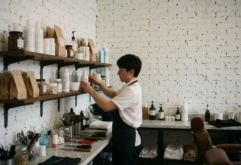 Un barbier range des articles sur une étagère en bois dans un salon avec murs de briques blanches, lumière naturelle et équipements visibles.