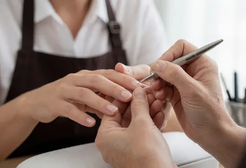 Premier plan des mains d'une cliente pendant qu'une manuciste professionnelle applique un soin avec un outil, sur fond flou et éclairage doux.