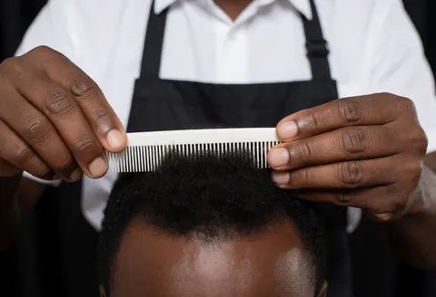 Un coiffeur professionnel mesure la longueur des cheveux d'un client avec une peigne blanche sur fond sombre, mettant en valeur la précision du service.