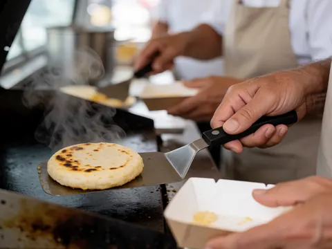 Des arepas en train de cuire sur une grille, avec de la vapeur s'élevant, préparées par des mains dans un contexte de food truck.