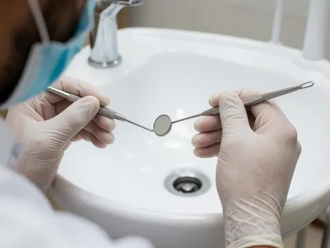 Mains d'un professionnel de l'odontologie ajustant un miroir et un explorateur dentaire sur un lavabo blanc, avec des gants et un masque dans un environnement clinique propre.