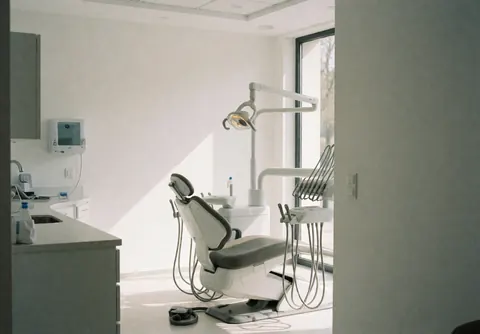Vue intérieure d'un cabinet dentaire contemporain avec chaise de traitement, éclairage chirurgical et murs blancs baignés de lumière naturelle.