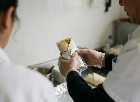Mains gantées en train de préparer un burrito chaud enveloppé dans du papier aluminium, avec de la vapeur s'élevant, sur un fond flou blanc.