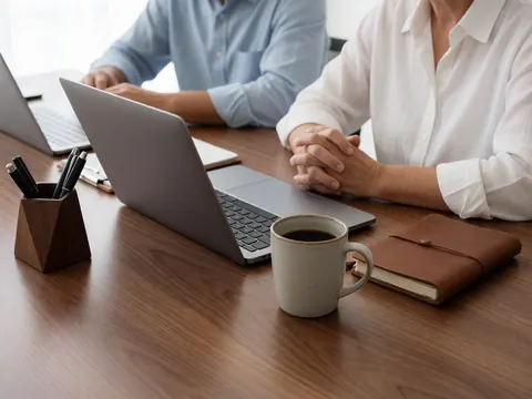 Deux personnes travaillent à une table en bois dans un environnement de coworking moderne, accompagnées d'un ordinateur portable, d'une tasse de café, d'un carnet et d'un porte-stylos.