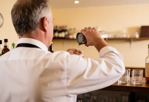 Un serveur professionnel sert une boisson derrière le comptoir d'un bar avec une ambiance chaleureuse et des murs clairs en arrière-plan flou.