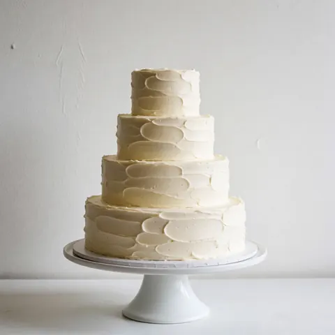 Un gâteau de mariage blanc minimaliste posé sur un piédestal, avec une texture de glaçage visible, sur fond blanc uni.