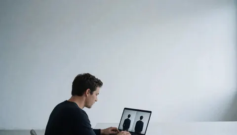 Un photographe adulte examine des images sur son laptop dans un studio minimaliste aux murs blancs et tons froids, avec de l'espace pour du texte.