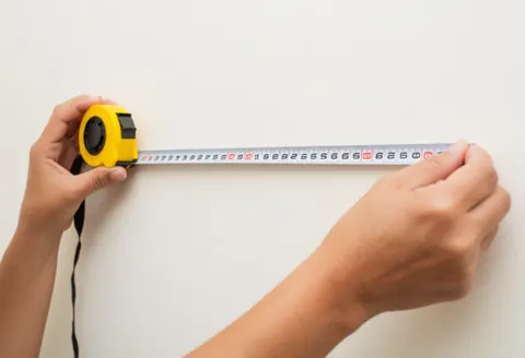 Des mains tenant un mètre ruban pour mesurer la largeur d'un mur blanc dans le cadre d'une évaluation immobilière ou de travaux de construction.