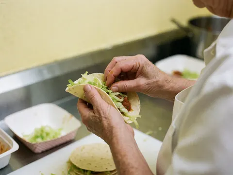 Des mains préparent des tacos en ajoutant de la laitue et de la sauce dans une cuisine aux murs jaunes, mettant en valeur les textures des ingrédients.