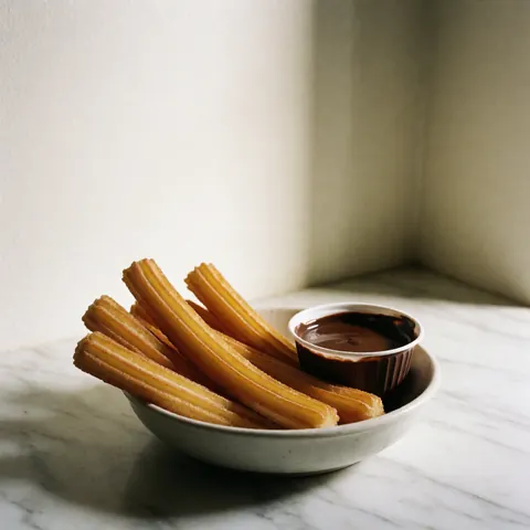 Des churros dorés saupoudrés de sucre sont disposés sur une assiette blanche, accompagnés d'un verre de chocolat chaud, le tout posé sur un plan de travail en marbre blanc baigné de lumière naturelle douce.