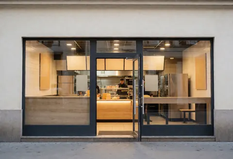 Façade moderne d'un restaurant avec une porte vitrée ouverte, intérieur lumineux et une personne travaillant au comptoir, ambiance chaleureuse et propre.