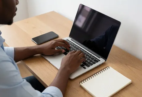 Une personne utilise un ordinateur portable posé sur un bureau en bois, accompagnée d'un téléphone et d'un cahet dans un environnement professionnel lumineux.