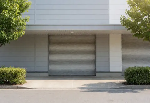 Façade d'un entrepôt logistique avec porte métallique enroulable, murs gris, arbres et arbustes dans une zone périurbaine. Éclairage naturel et clair.