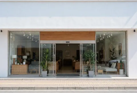 Vue extérieure d'une devanture de magasin de mobilier moderne avec grandes fenêtres en verre, portes d'entrée et plantes décoratives sur le trottoir.