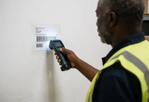 Un opérateur en gilet de sécurité scanne un code-barres sur un mur blanc d'entrepôt, illustrant la précision logistique et technologique.