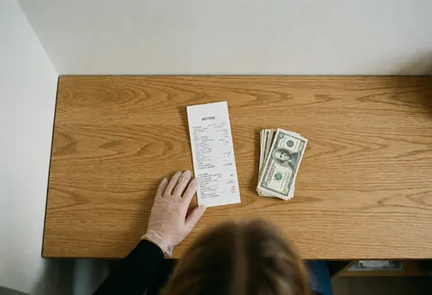 Caisse enregistreuse accompagnée d'un reçu et de billets de dollar posés sur une table en bois clair, évoquant une scène de paiement.
