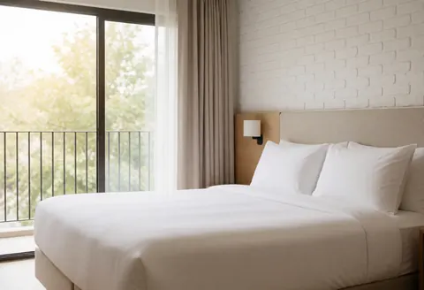 Chambre d'hôtel moderne avec un grand lit, un mur en briques blanches, une fenêtre offrant une vue sur le balcon et une lumière naturelle abondante.