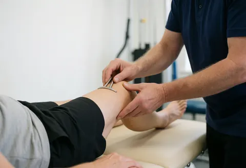 Physiothérapeute effectuant un traitement sur le genou d'un patient allongé sur une table de consultation dans un environnement clinique professionnel et lumineux.