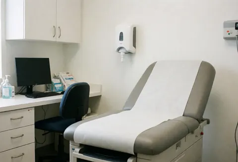 Vue intérieure d'un cabinet médical moderne avec une table d'examen, un bureau équipé d'un ordinateur et des équipements de désinfection.