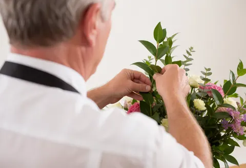 Un homme de dos arrange un bouquet de fleurs composé de roses et de feuillage vert dans un studio éclairé, illustrant le travail en floristerie.