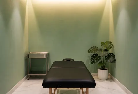 Intérieur d'une salle de massage au style épuré avec des murs peints en vert, un sol en marbre blanc brillant et une table de massage noire. Une plante monstera apporte une touche naturelle sous un éclairage chaleureux.