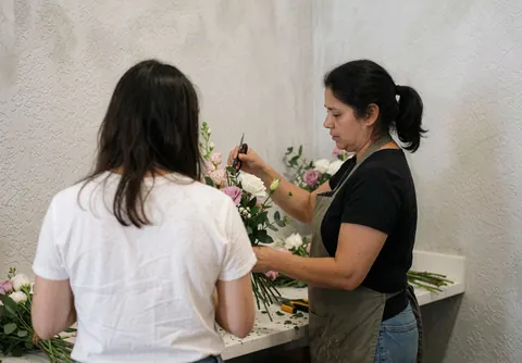 Une fleuriste travaille sur un bouquet avec une cliente dans son atelier, utilisant des ciseaux et des fleurs sur une table devant un mur gris.