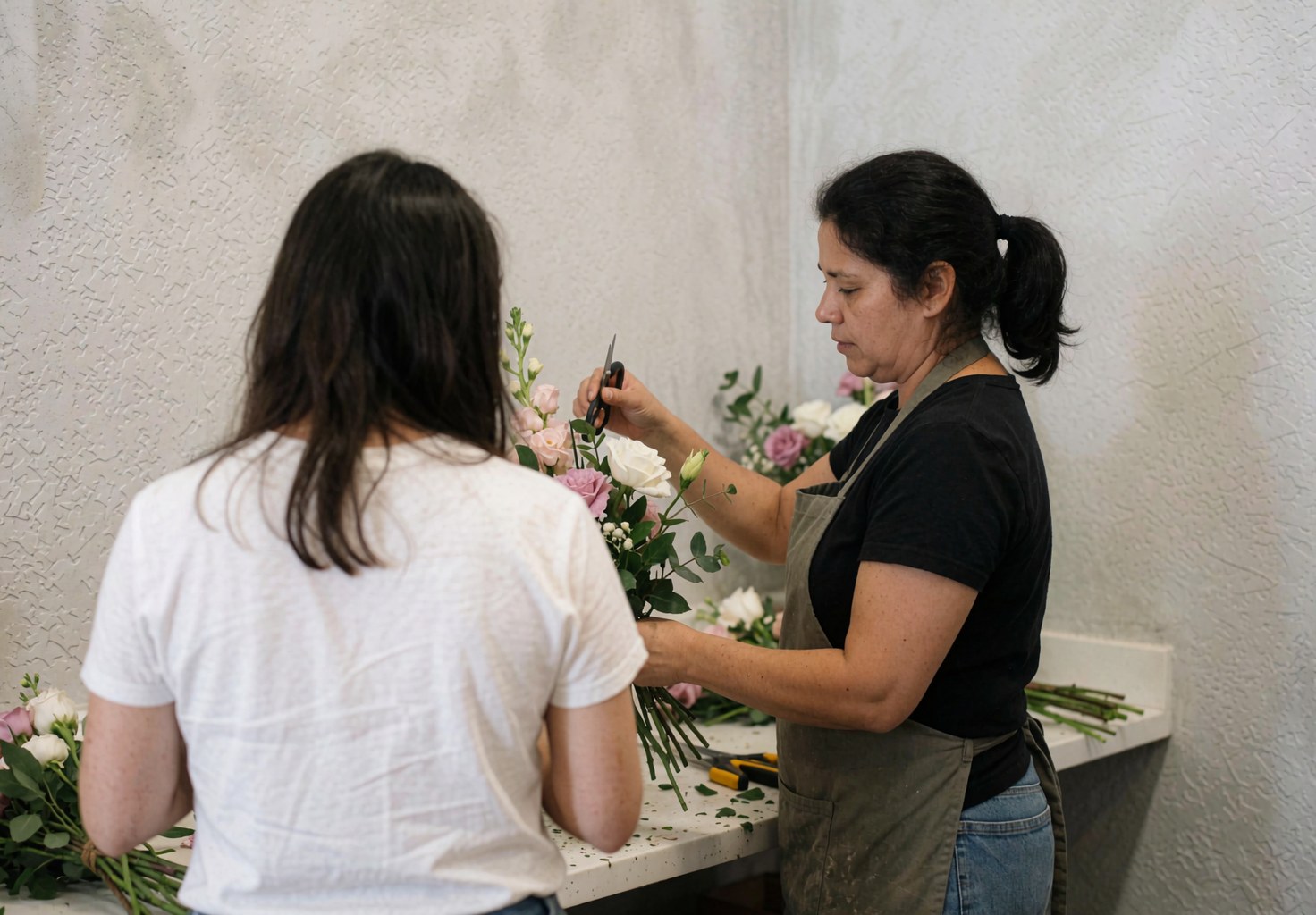 Une fleuriste travaille sur un bouquet avec une cliente dans son atelier, utilisant des ciseaux et des fleurs sur une table devant un mur gris.