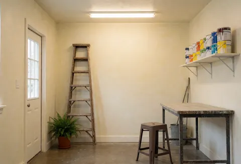 Atelier de peinture vide aux murs clairs, équipé d'une échelle en bois, de peintures sur étagère, d'une table de travail et d'une chaise plastique.