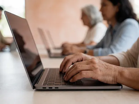 Gros plan sur des mains qui écrivent sur le clavier d'un ordinateur portable posé sur une table en bois, avec un écran visible et un fond flou.